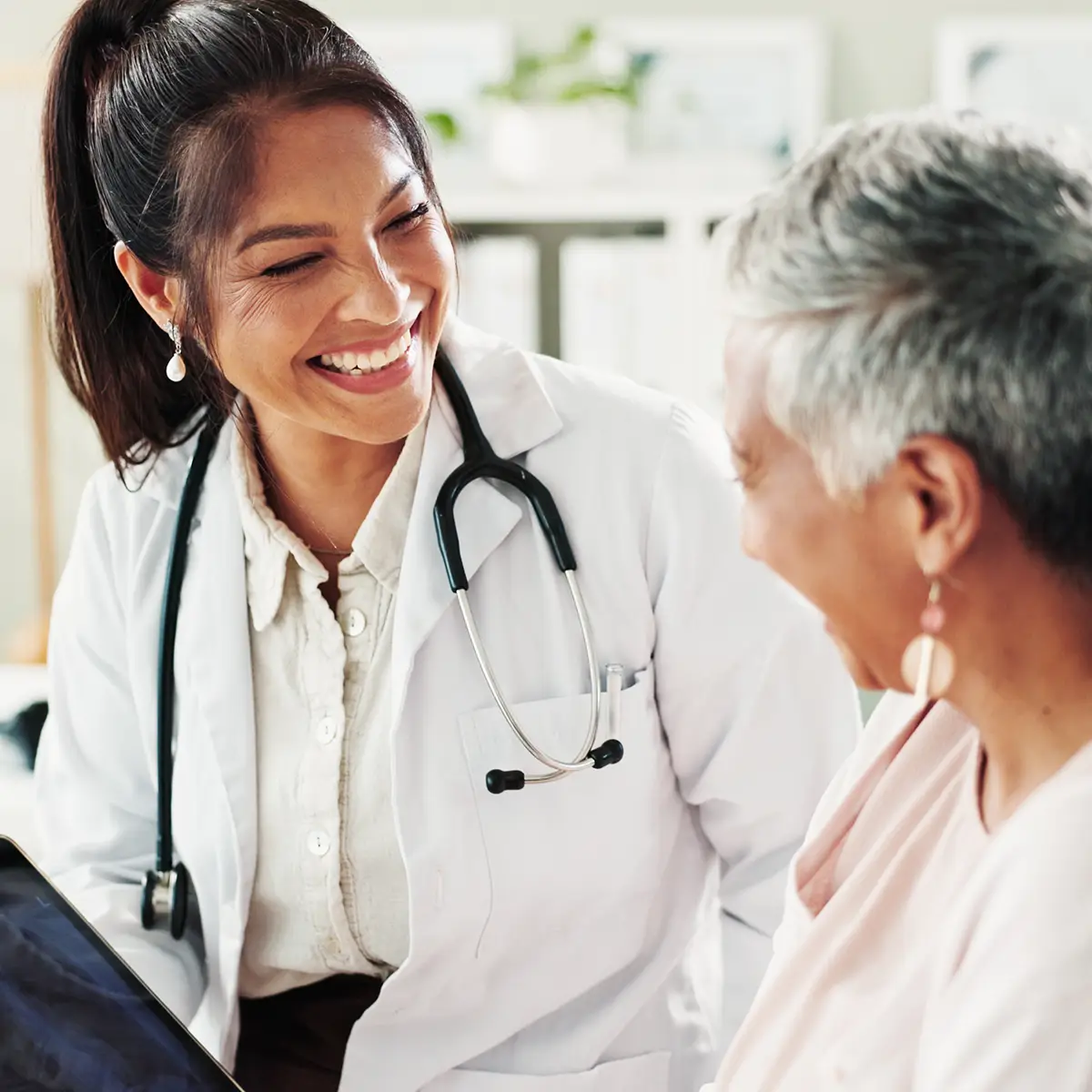A physician in a white coat and stethoscope smiling with an older patient during a consultation